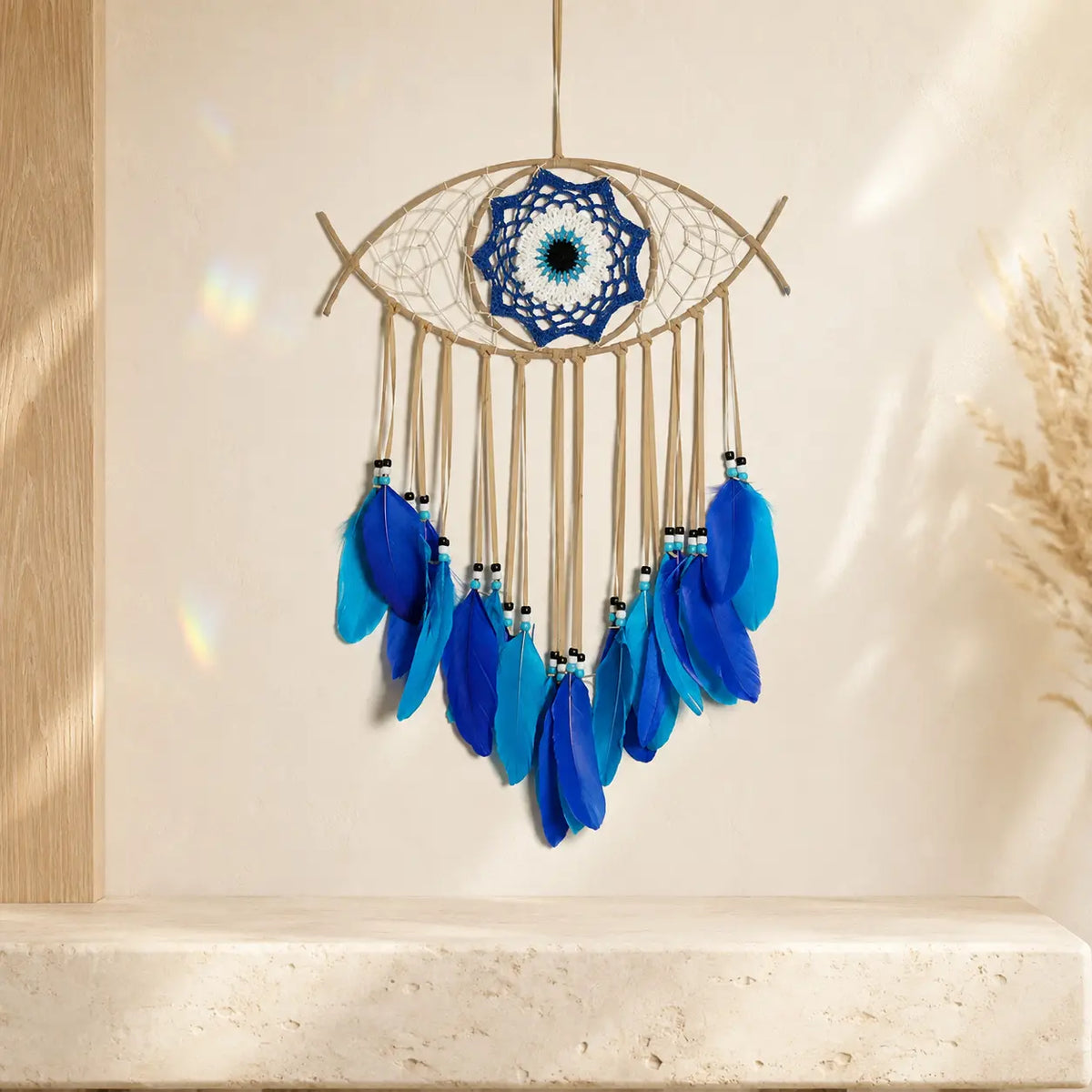 An evil eye dreamcatcher wall hanging suspended from a wooden branch, featuring a blue and white webbed center, tan string webbing, and multiple dangling blue feathers, displayed against a warm beige wall with soft sunlight and shadows.