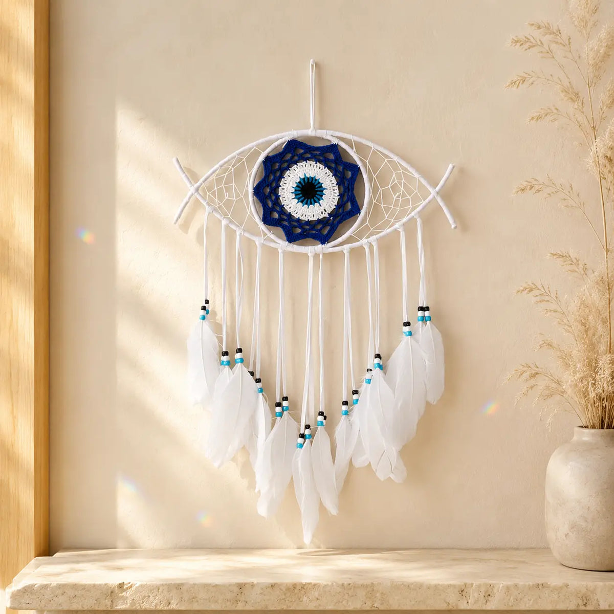 White Evil Eye Dream Catcher wall hanging with a blue and white evil eye center, suspended on a beige wall above a stone ledge with dried grass in a vase.