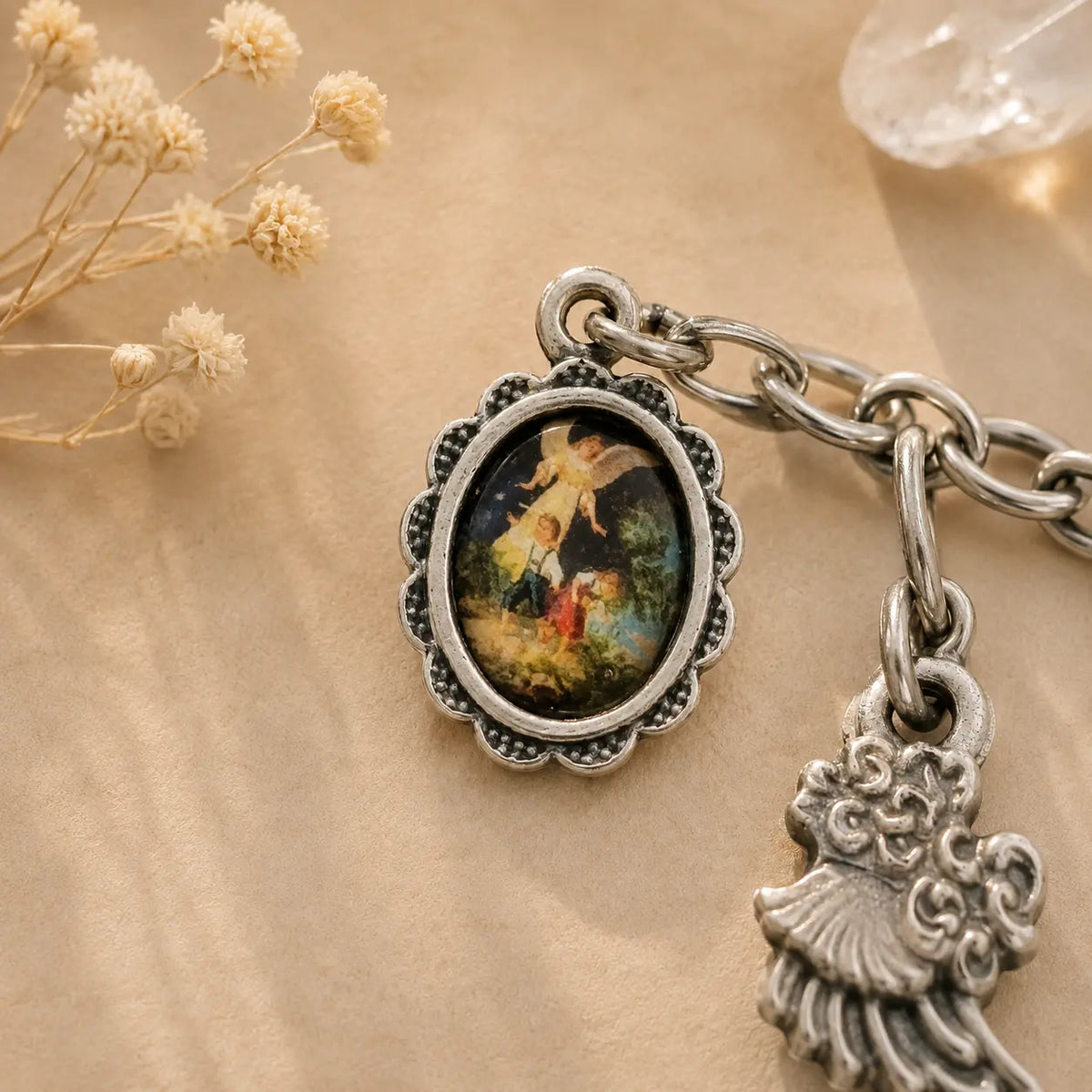 An antique-style silver charm bracelet featuring an oval pendant with a miniature classical painting of an angel and a child, alongside a dangling angel figure charm, displayed on a beige fabric background with dried flowers.