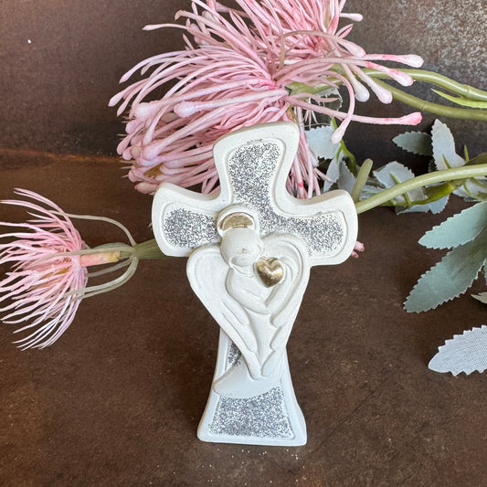 Decorative cross with angel holding a heart, surrounded by pink flowers on a brown surface