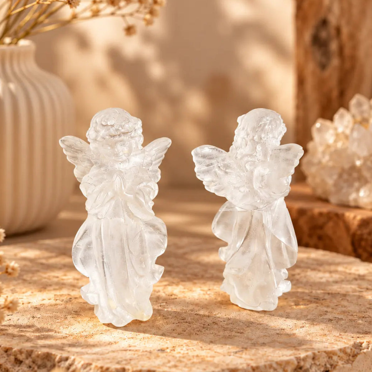 Two small clear quartz angel figurines standing on a round stone slab, styled with dried flowers and a crystal cluster in warm natural light. Approximately 10cm for clarity, peace and spiritual protection