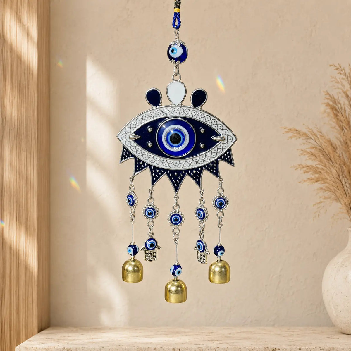 Blue Evil Eye Wind Chime/Suncatcher with two Hamsa hand charms and three gold-tone bells hanging against a warm cream wall