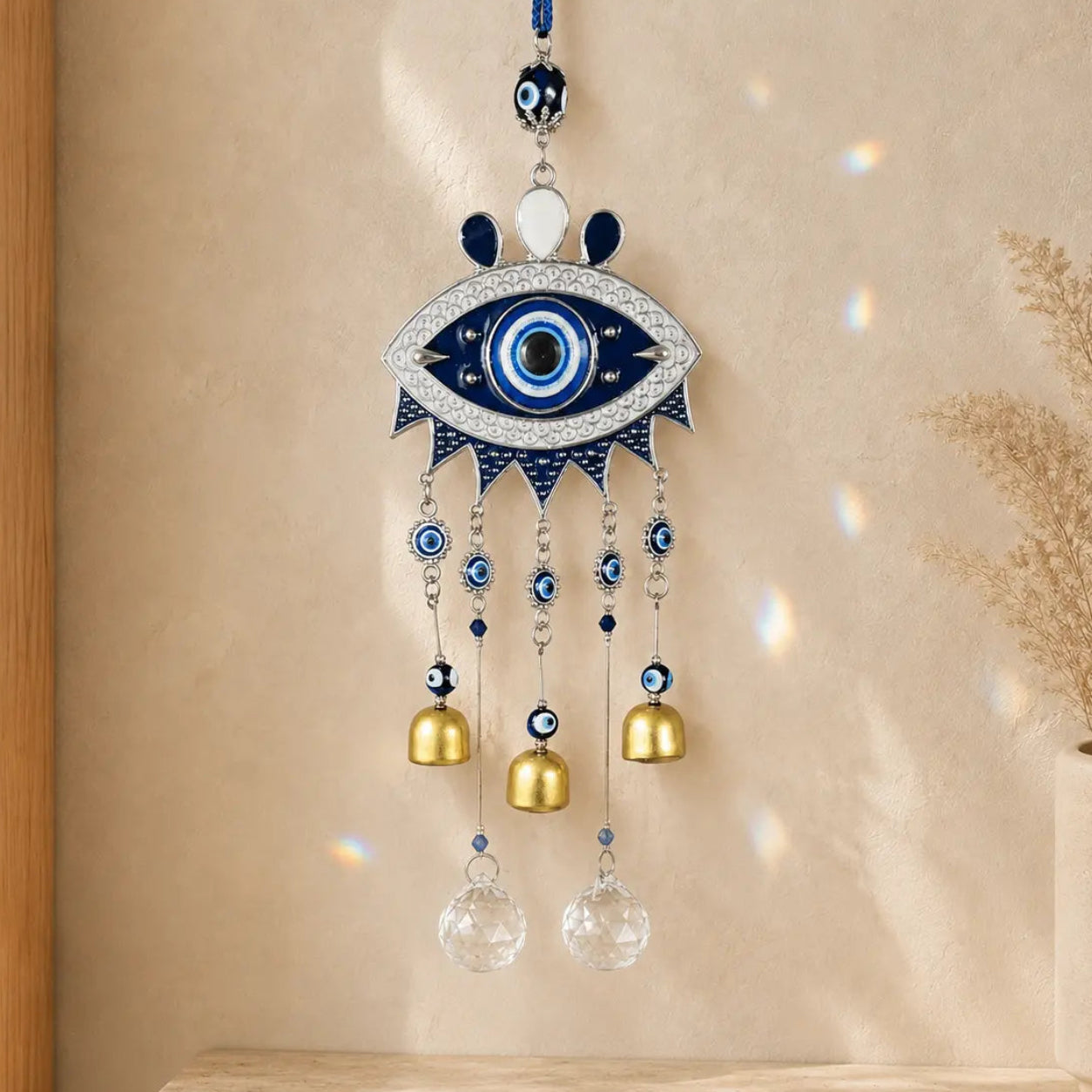 Blue Evil Eye wind chime with three gold-tone bells and two clear faceted suncatcher drops hanging against a warm cream wall