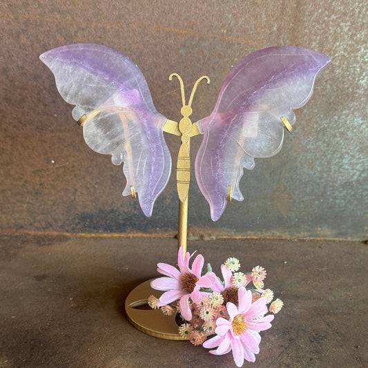 Decorative butterfly with purple wings and gold body on a stand with pink flowers against a brown background