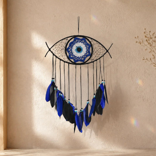 Handmade_Evil_Eye_Dream_Catcher_Black wall hanging shaped like an eye, with a blue and white circular center and dangling black, blue, and navy feathers, displayed against a warm beige textured wall on a stone ledge.
