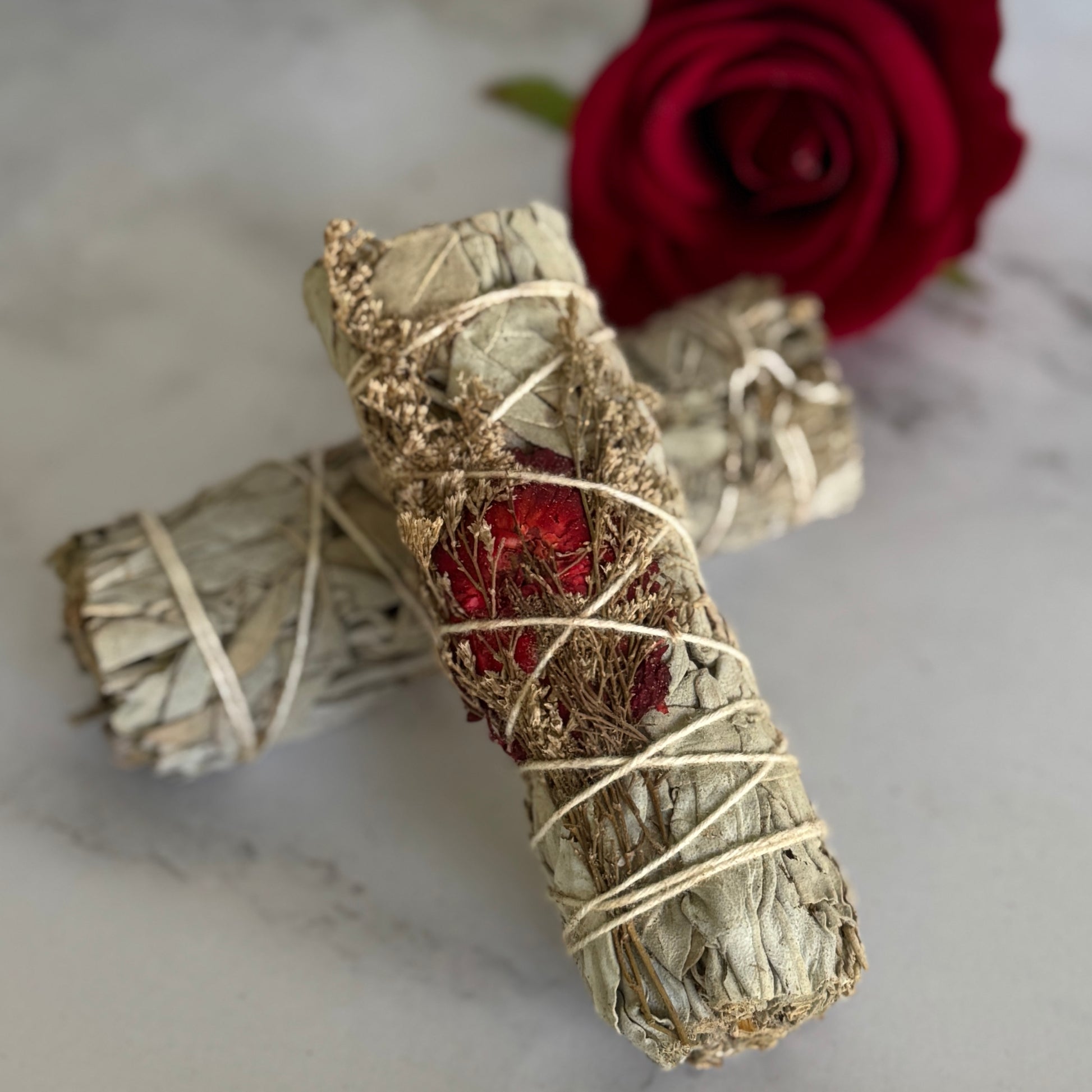 Two white sage smudge sticks bound with string, one with a red flower embedded in it, displayed on a marble surface with a red rose in the background.