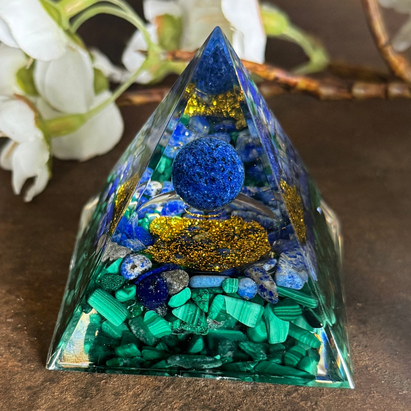 A triangular Orgonite Pyramid Malachite & Lapis Lazuli with Sodalite Sphere, featuring a collection of healing stones inside, with a blue spherical stone at the top, placed on a wooden surface with white flowers in the background.