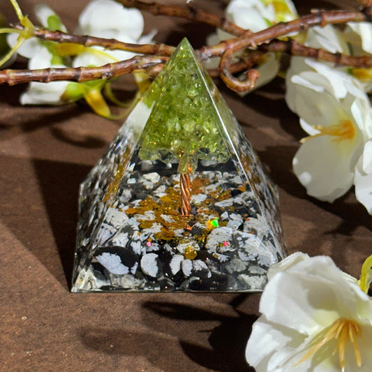 An Orgonite Pyramid with Peridot Tree placed on a wooden surface with white flowers around it. The pyramid is made of black and white materials with green peridot leaves and copper elements.