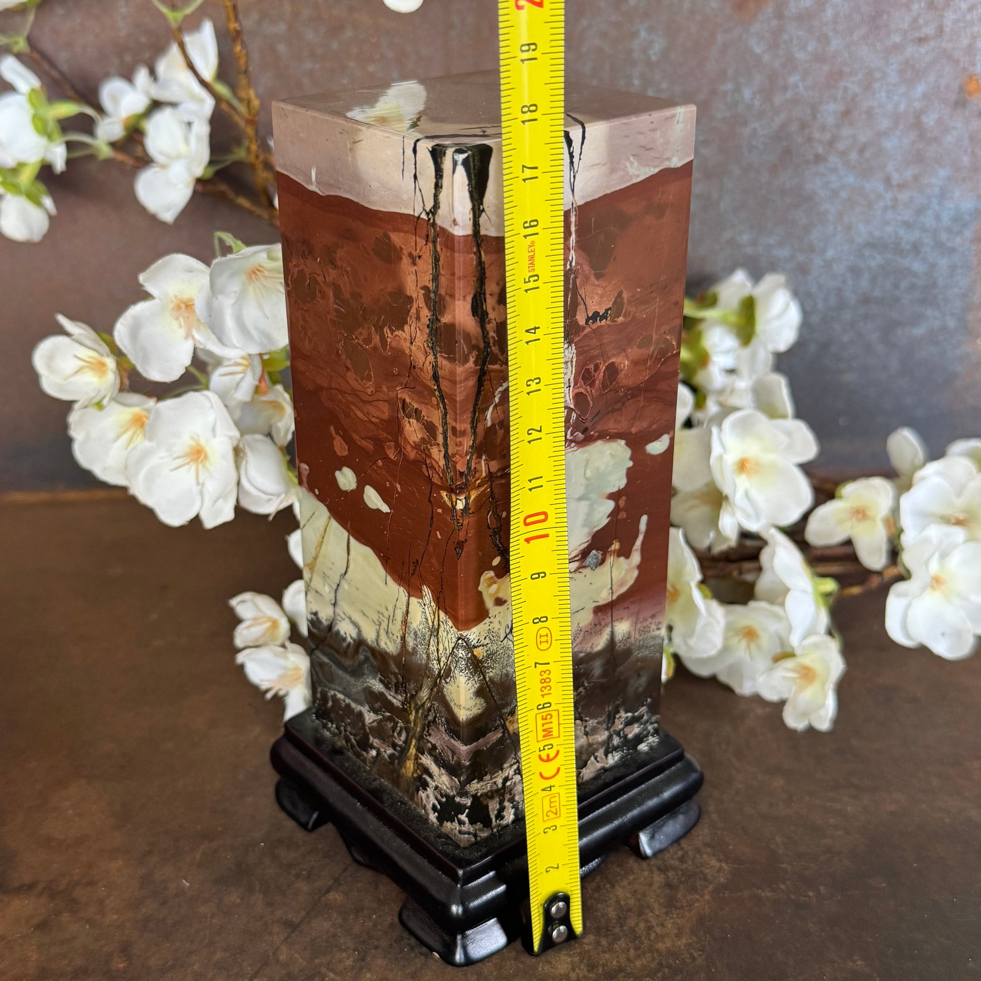 Picture Jasper crystal on timber stand for grounding energy, calm, emotional balance and spiritual décor statement piece.a measuring tape for scale, set against a floral background.