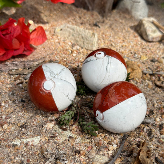 A novelty Pokémon crystal ball featuring red jasper and howlite, placed on a bed of gravel with a few dried leaves scattered around.