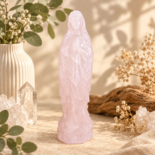 A pink rose quartz statue of the Virgin Mary stands centered on a beige surface, surrounded by dried flowers, green leaves, clear crystal clusters, and a ribbed cream vase in a warm, sunlit setting.