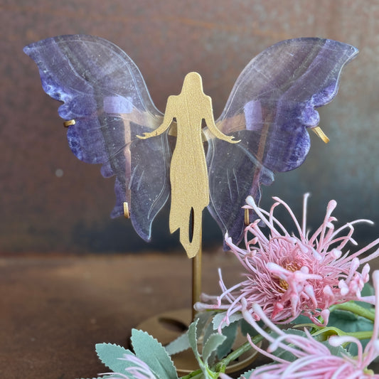 A crystal piece resembling wings attached to a stand, with a golden angel figure in the center, displayed alongside pink flowers.